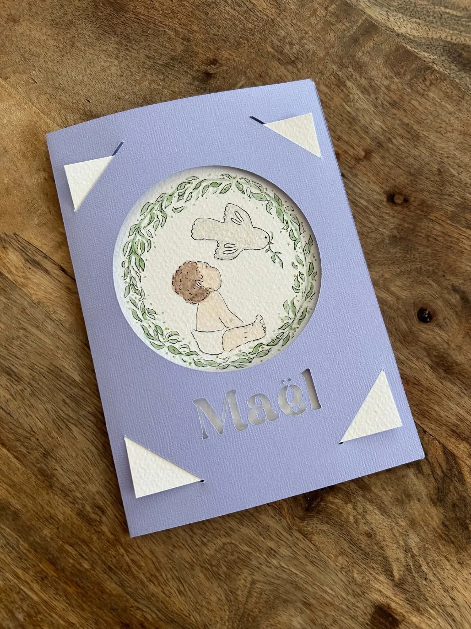 Carte de vœux de Noël avec un dessin d'un enfant tenant un oiseau et une branche de gui, entourée de feuilles vertes, posée sur une table en bois.