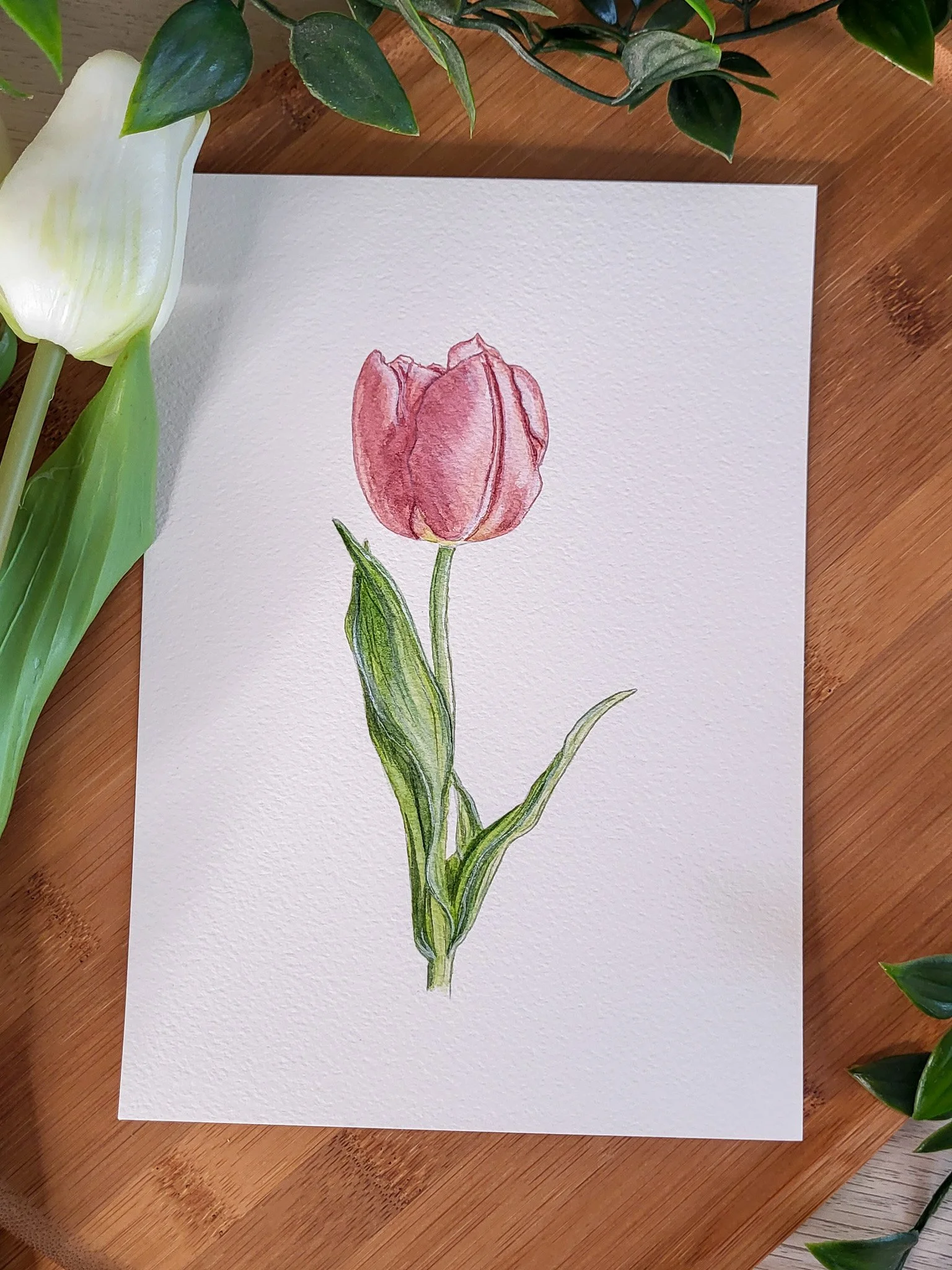 Une peinture d'une tulipe rose sur papier blanc, posée sur une table en bois avec des feuilles vertes autour.