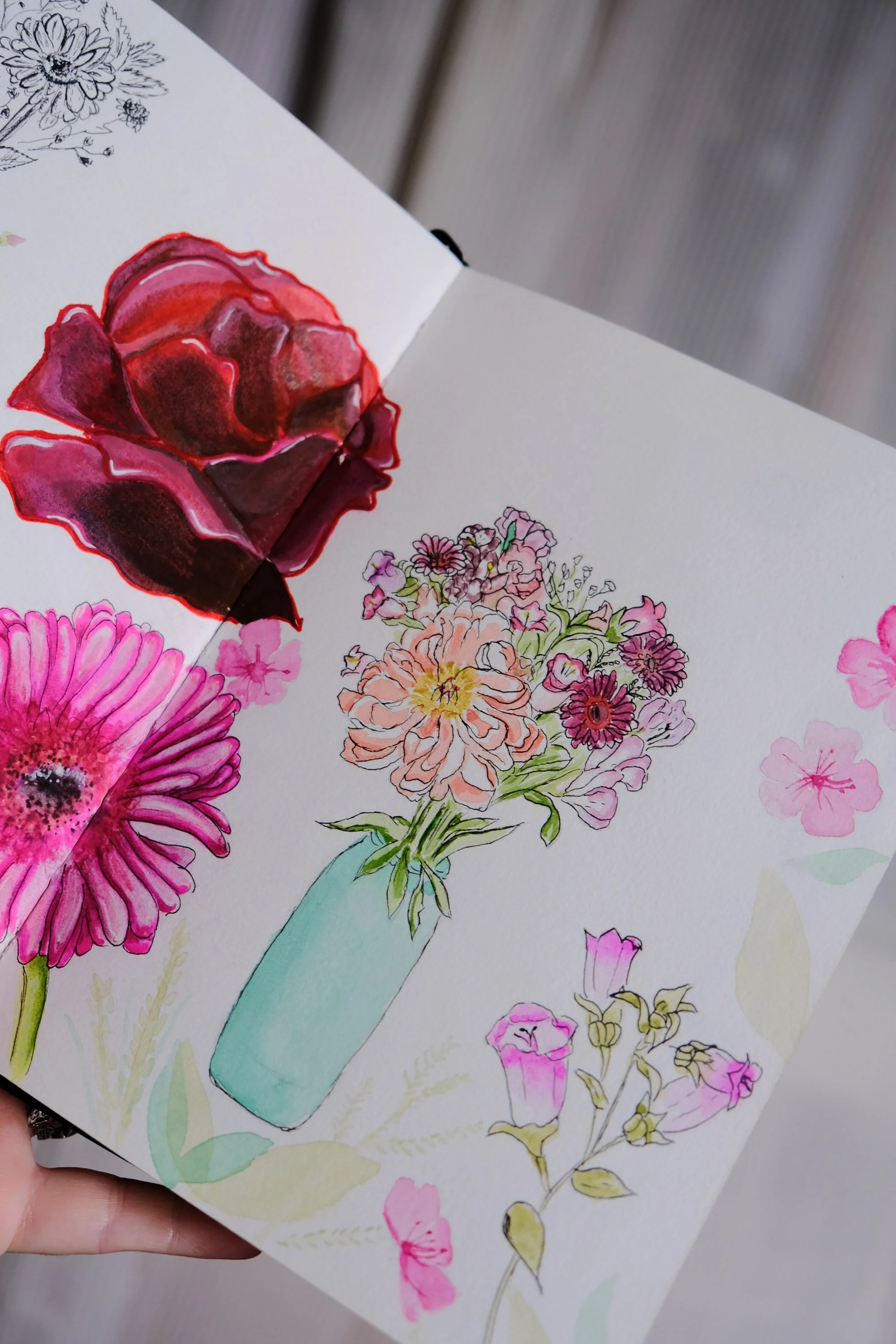 Une main tient un carnet où sont dessinées et peintes des fleurs colorées et un bouquet dans un vase en turquoise.