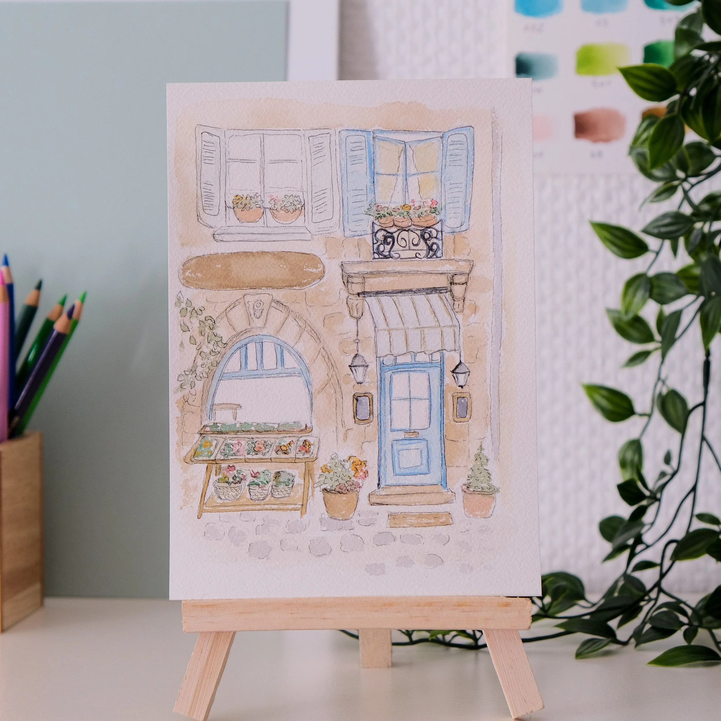 Une aquarelle représentant une fenêtre avec volets bleus, des pots de fleurs sur un rebord et une table avec un tapis