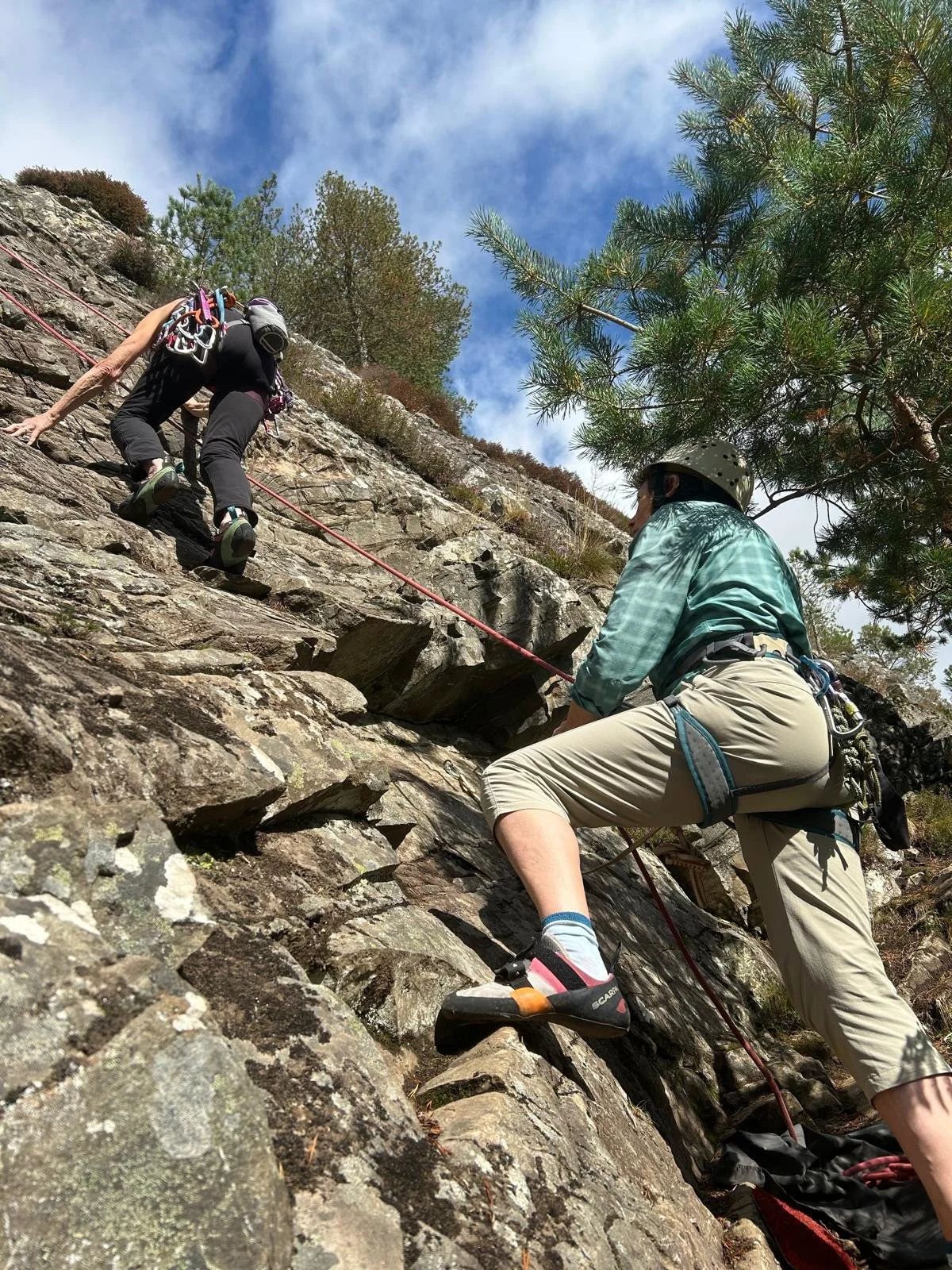 Two people rock climbing on a rugged, steep mountainside with climb gear, trees, and blue sky visible.