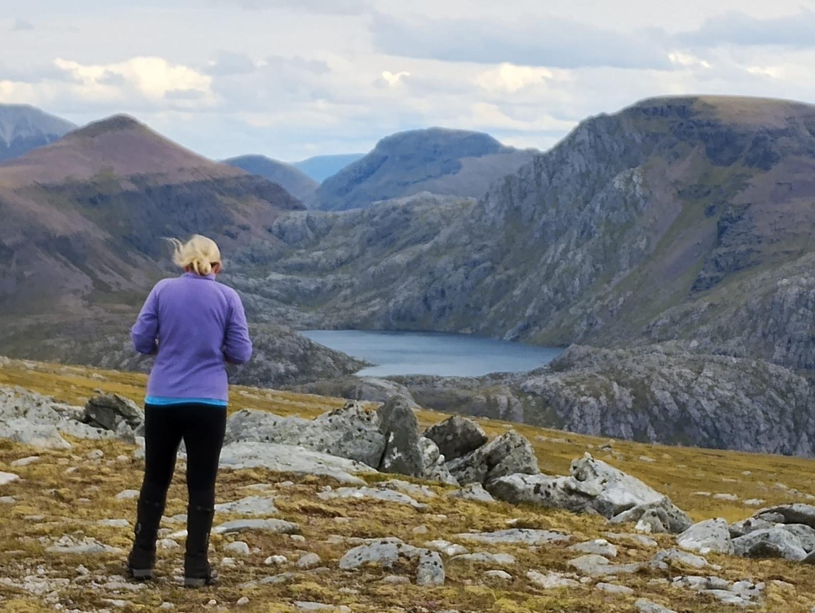 Person with blonde hair in a purple jacket standing on rocky terrain, overlooking a mountain valley with a lake and rolling hills under a partly cloudy sky.