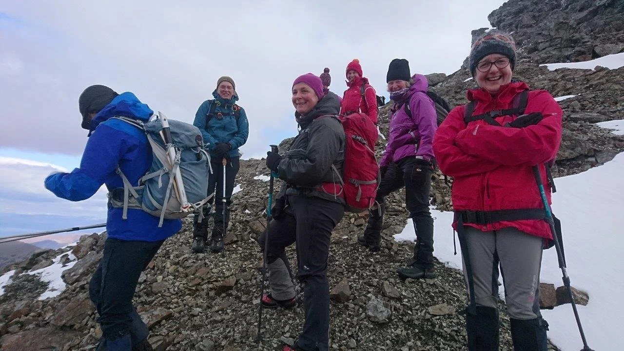 Group of six hikers wearing winter gear on a rocky mountain trail with snow, cloudy sky in the background.