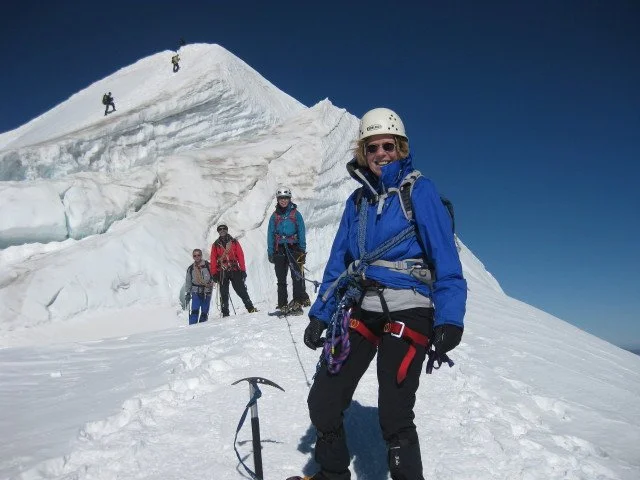 Group of five mountaineers in snow gear on a snowy mountain with a large ice formation, one woman in the foreground smiling, others climbing or standing in the background.