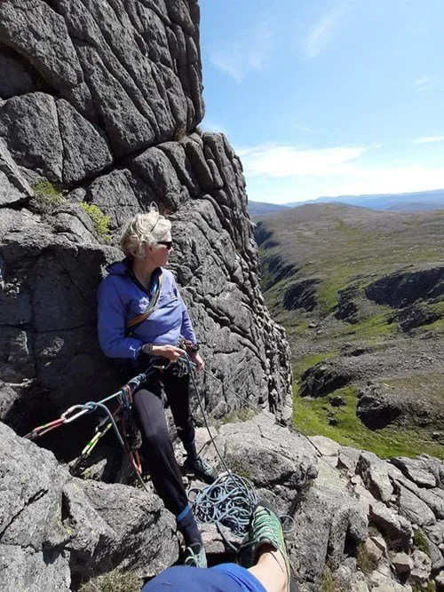 A woman sitting on rocks on a mountainside, wearing outdoor gear and sunglasses, with climbing equipment nearby, overlooking a green valley under a blue sky.