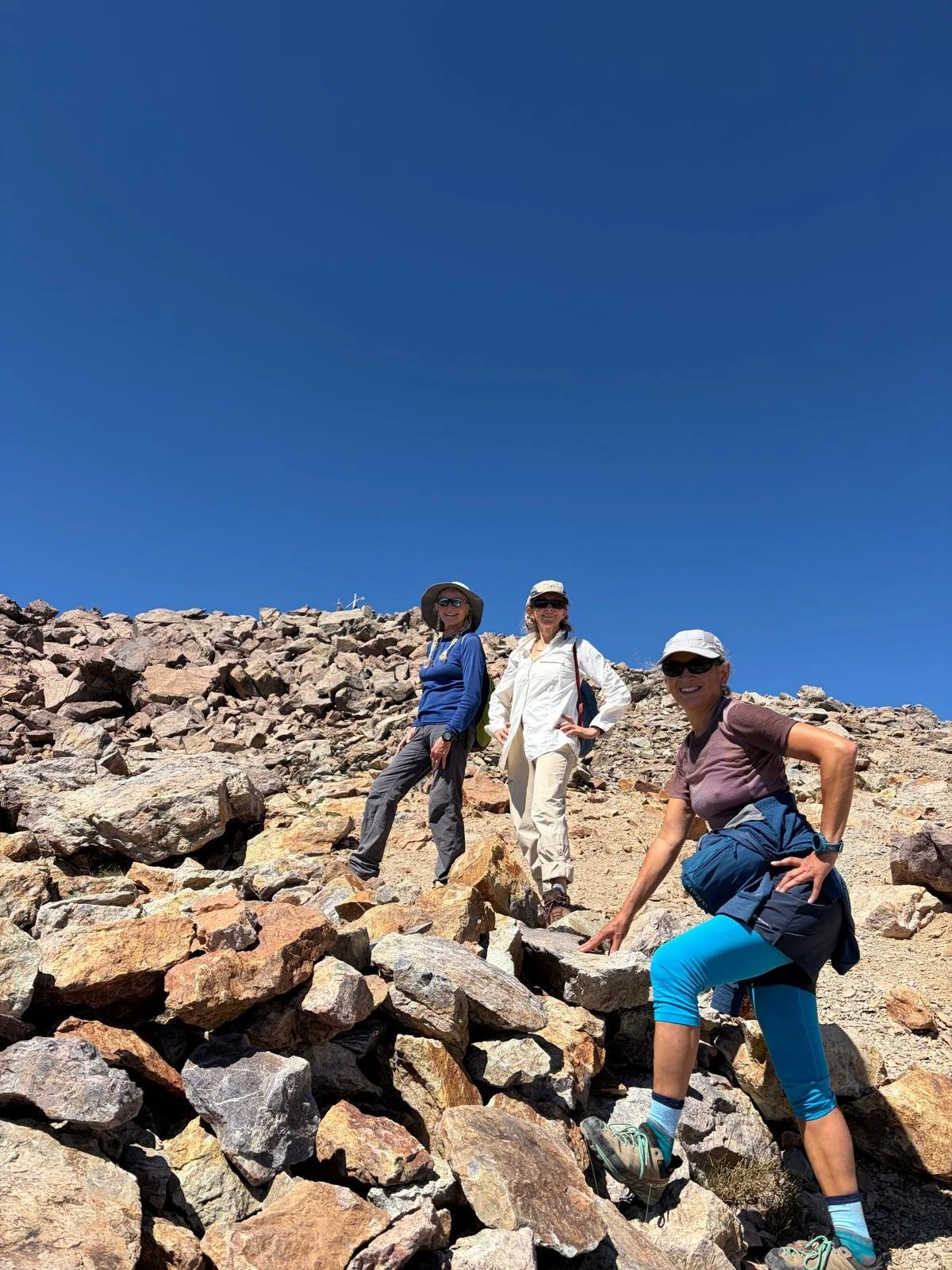 Three women hiking on rocky terrain under a clear blue sky, wearing hats, sunglasses, and outdoor clothing.
