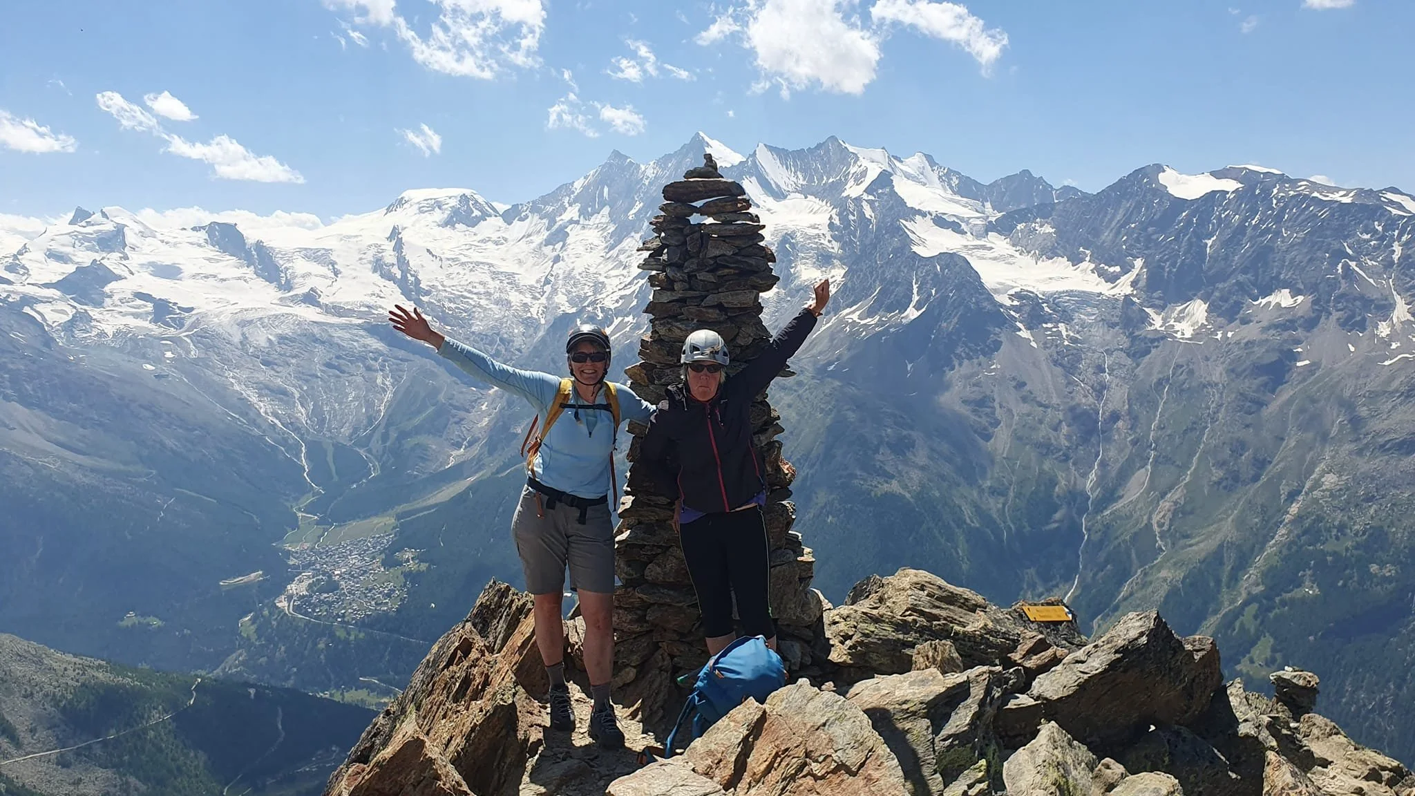 Two hikers posing on a mountain summit near a stone cairn with a snowy mountain range in the background during daytime.