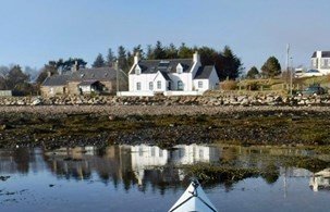 A coastal scene with houses along the shoreline and a reflection in the water, with a boat's bow visible in the foreground