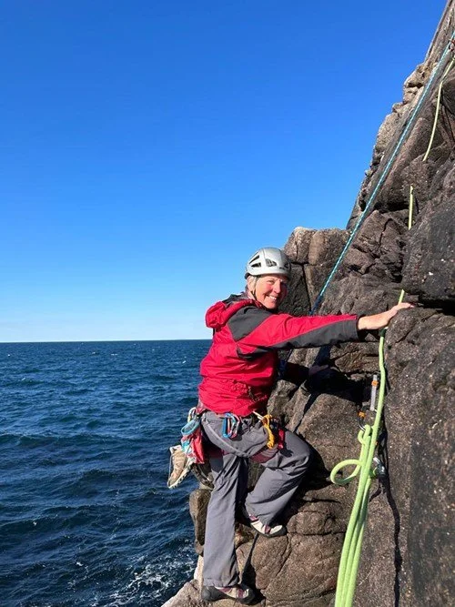 A woman in a red jacket and climbing gear ascending a rocky cliff by the ocean.