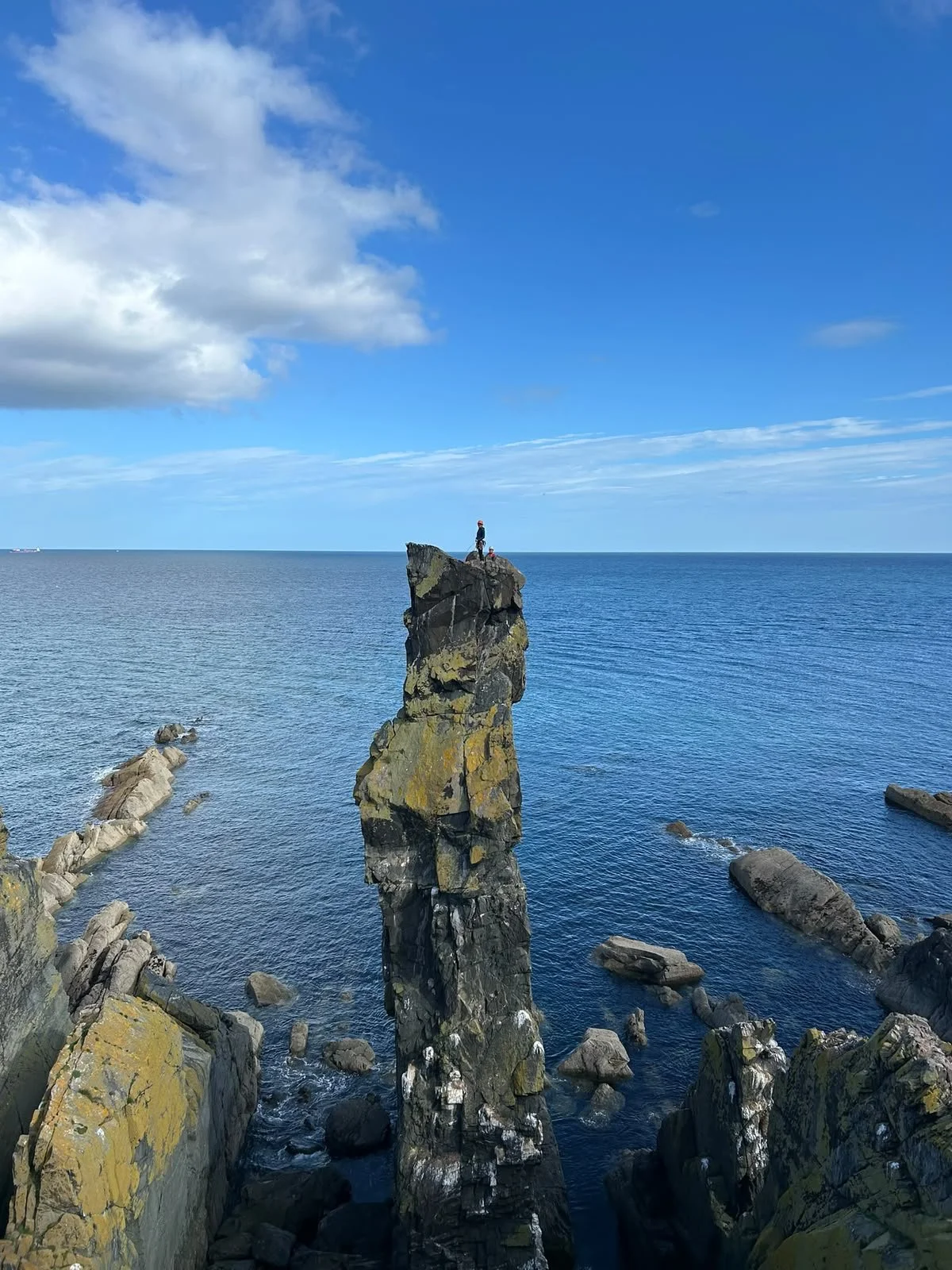A tall, narrow rock formation rising out of the ocean with two people standing at the top, surrounded by smaller rocks and a blue sky with scattered clouds.