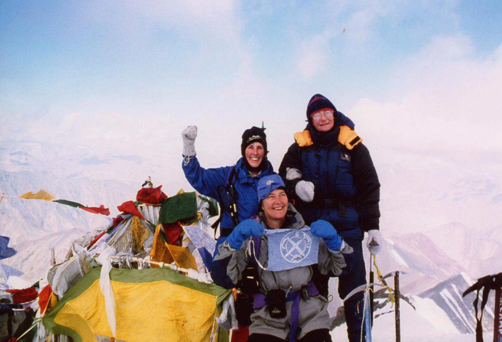 Three mountaineers on a snowy mountain summit, one holding a flag, celebrating victory, surrounded by prayer flags and icy peaks, overcast sky.