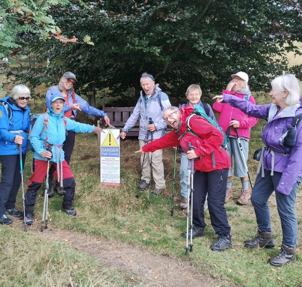 Group of senior hikers in outdoor gear standing around a warning sign that says 'Danger Path Closed' in a wooded area, smiling and pointing at the sign.
