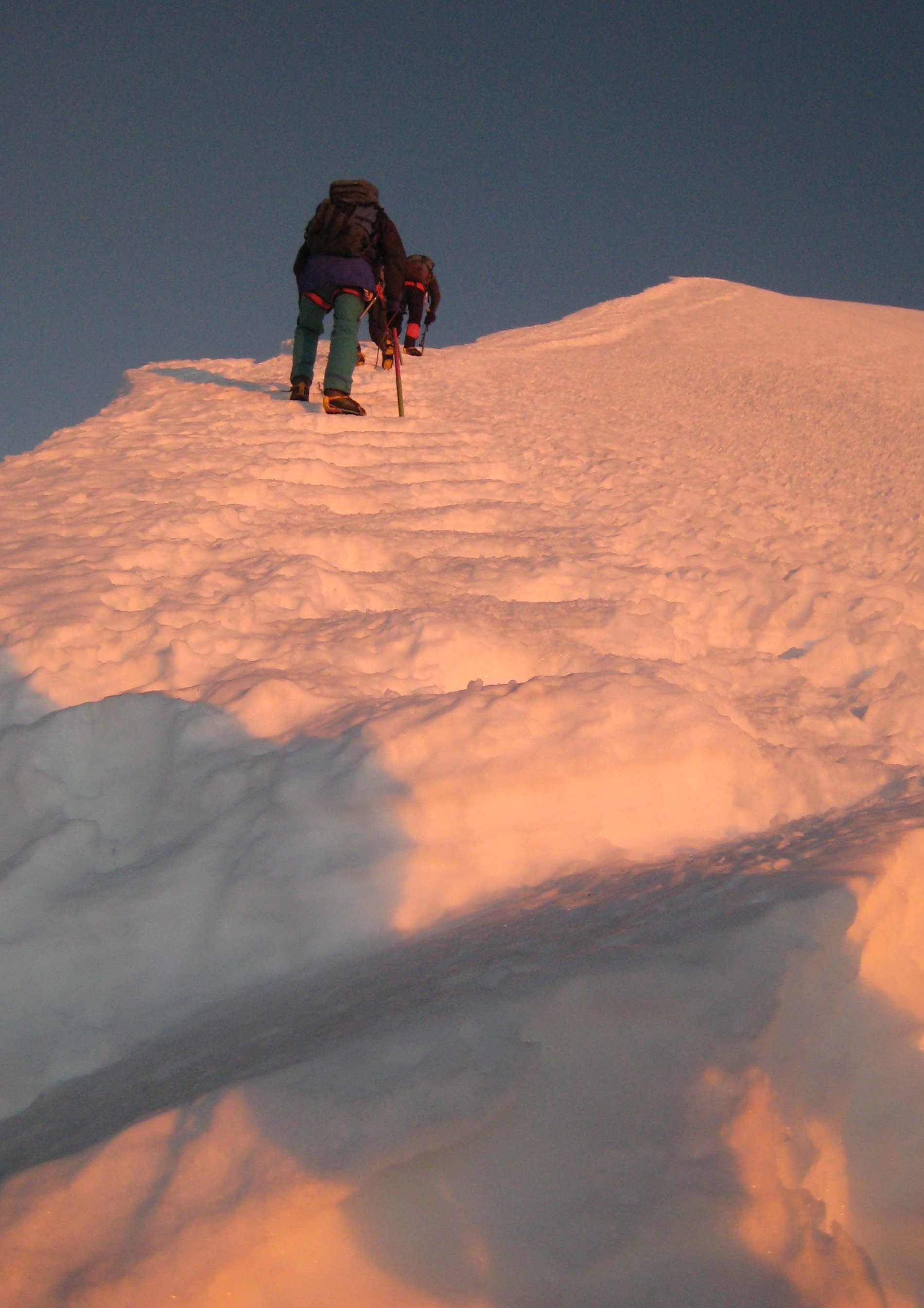 A group of mountaineers climbing a snow-covered peak during sunset or sunrise, with the sky displaying a warm glow.