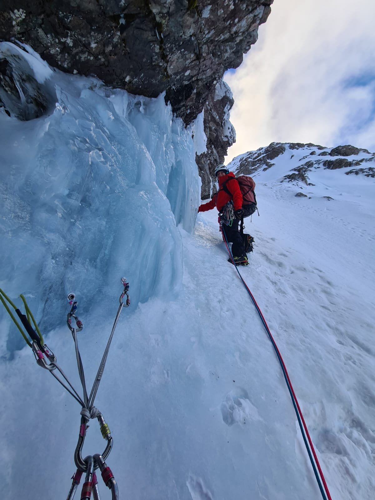 A climber dressed in red gear ascending an ice wall with ice axes and safety ropes in a snowy mountain environment.