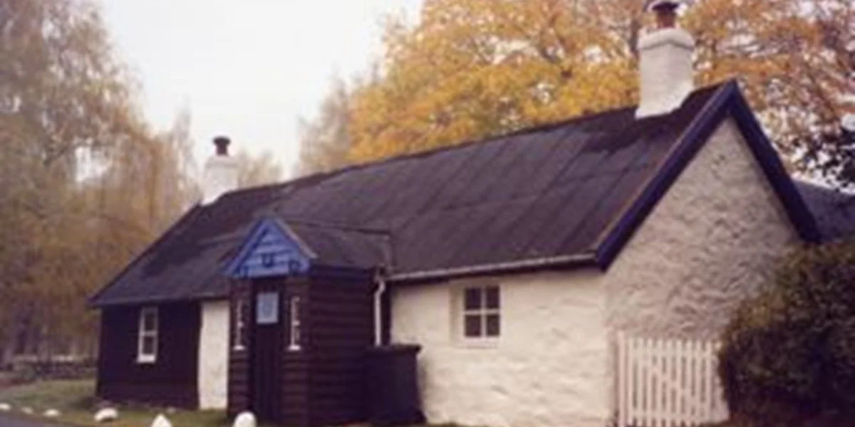 A small house with a mixed exterior of dark brown wood and white stone, a gabled roof, and a white chimney. The house is surrounded by trees with fall-colored leaves.