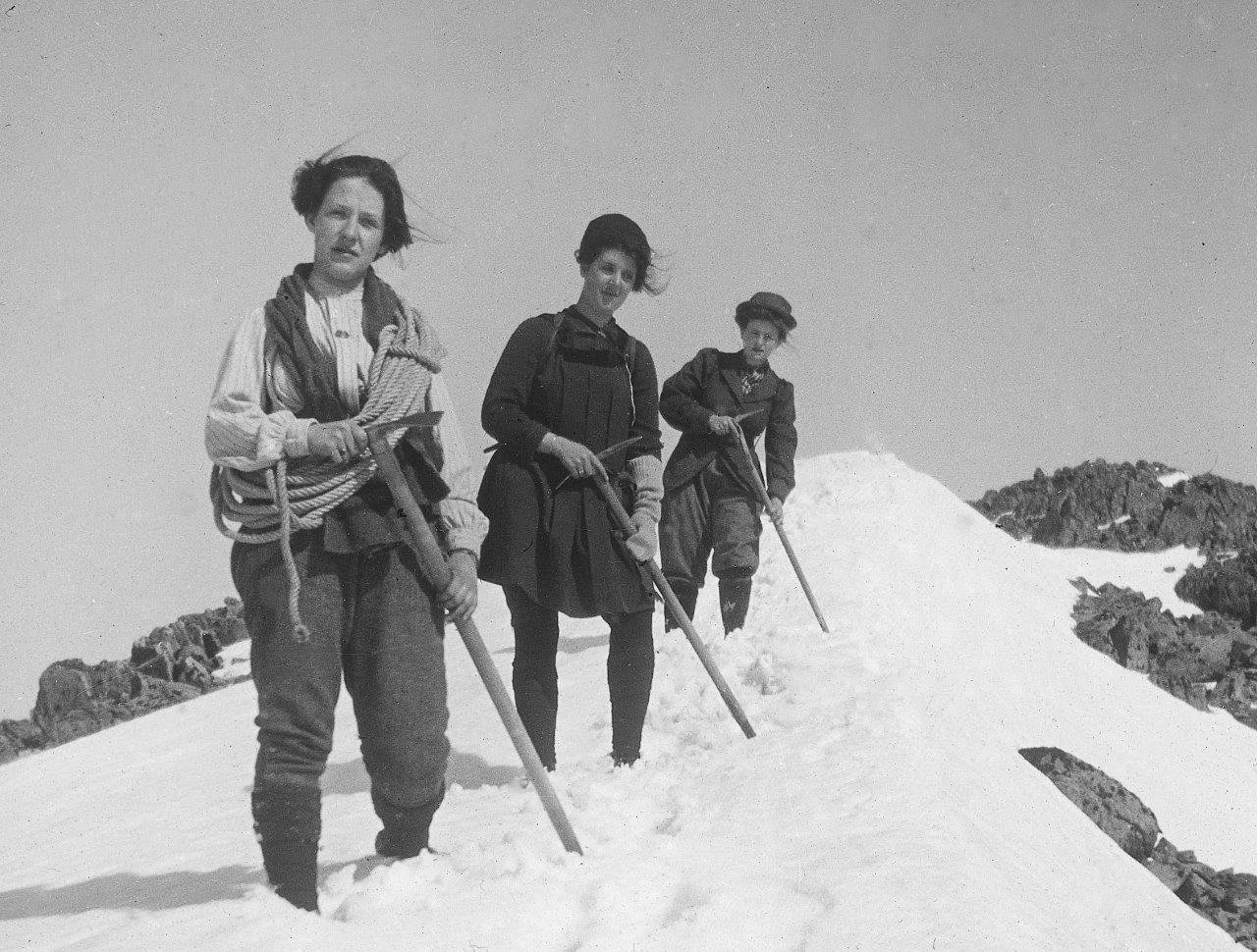 Three women standing in snow on a mountain, holding ice axes, dressed in early 20th-century outdoor clothing.