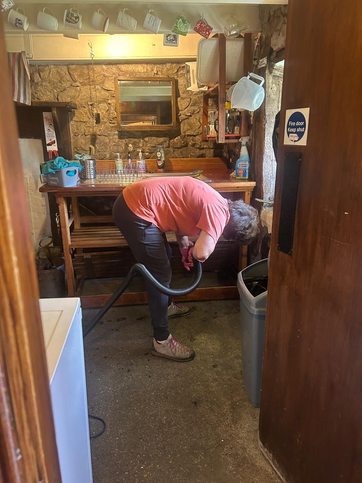 Person cleaning the floor in a rustic kitchen with stone walls, wooden furniture, hanging mugs, and cleaning supplies.