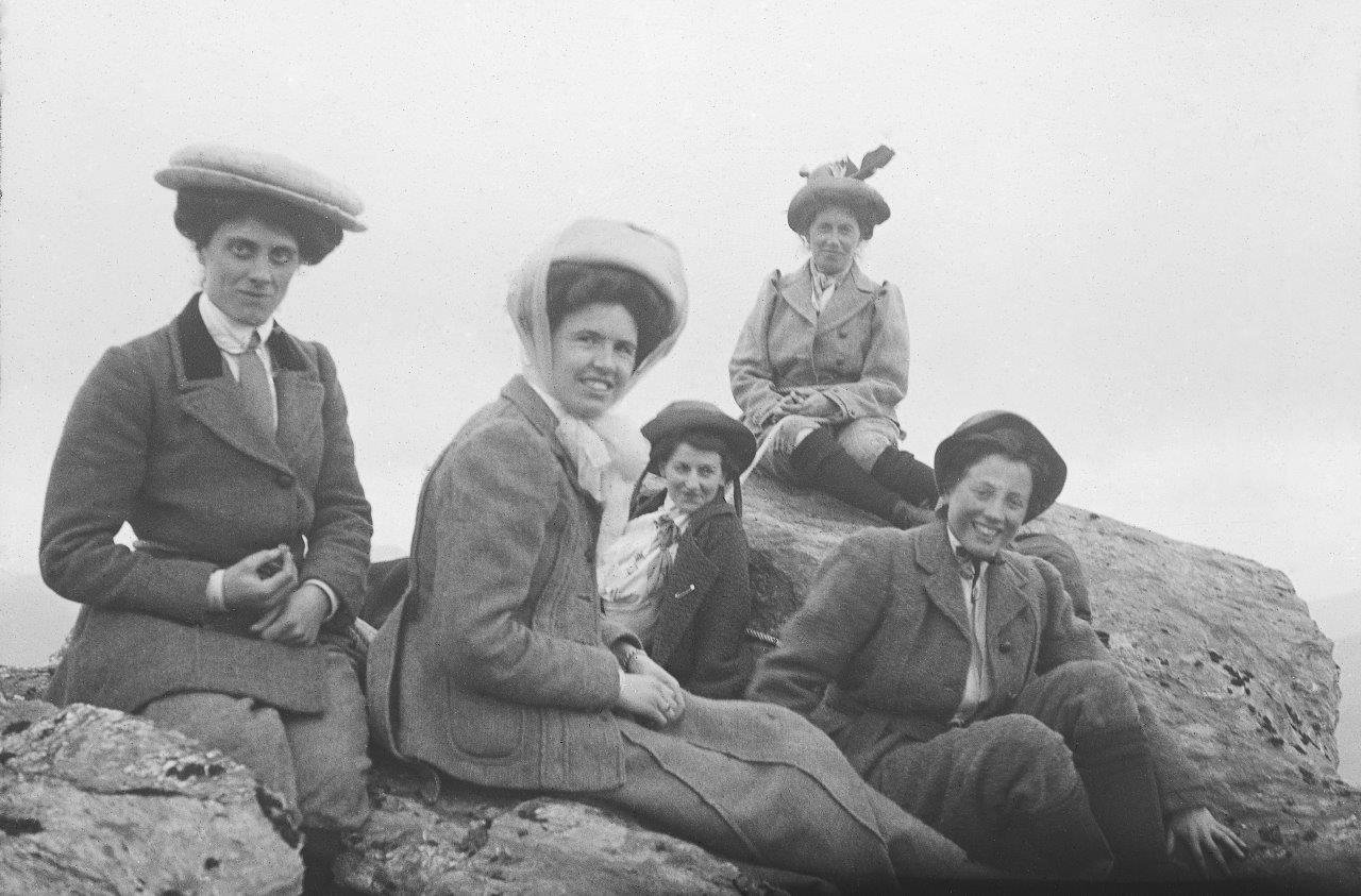 Five women in early 20th-century clothing sitting and standing on rocks outdoors, wearing hats.