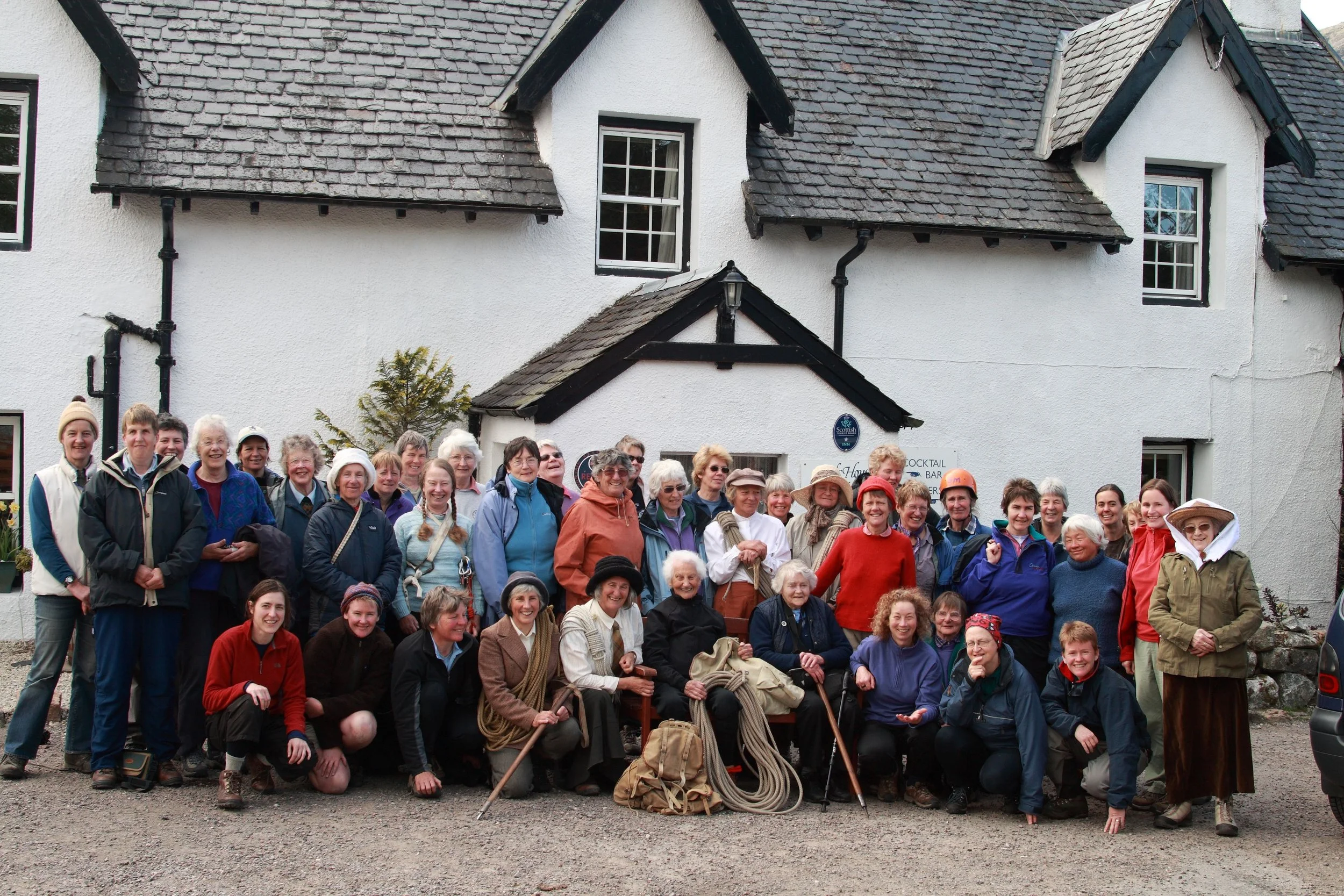 A large group of smiling people posing for a photo outside in front of a white building with dark roof shingles. The group includes men and women of various ages, some wearing outdoor gear and hats.