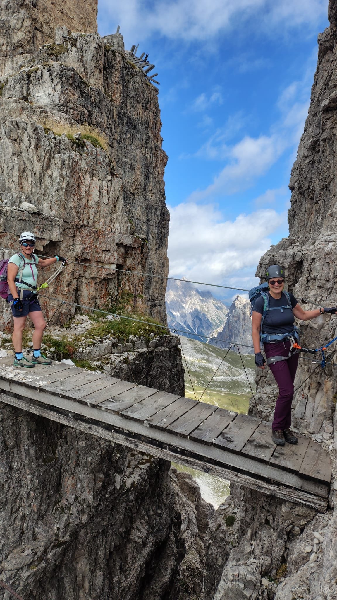 Two women with helmets and backpacks walking on a narrow wooden bridge between two rocky mountain cliffs, with a scenic mountain landscape in the background.