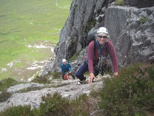 Two people climbing a steep rocky outdoor trail, wearing helmets and climbing gear, with a grassy landscape in the background.