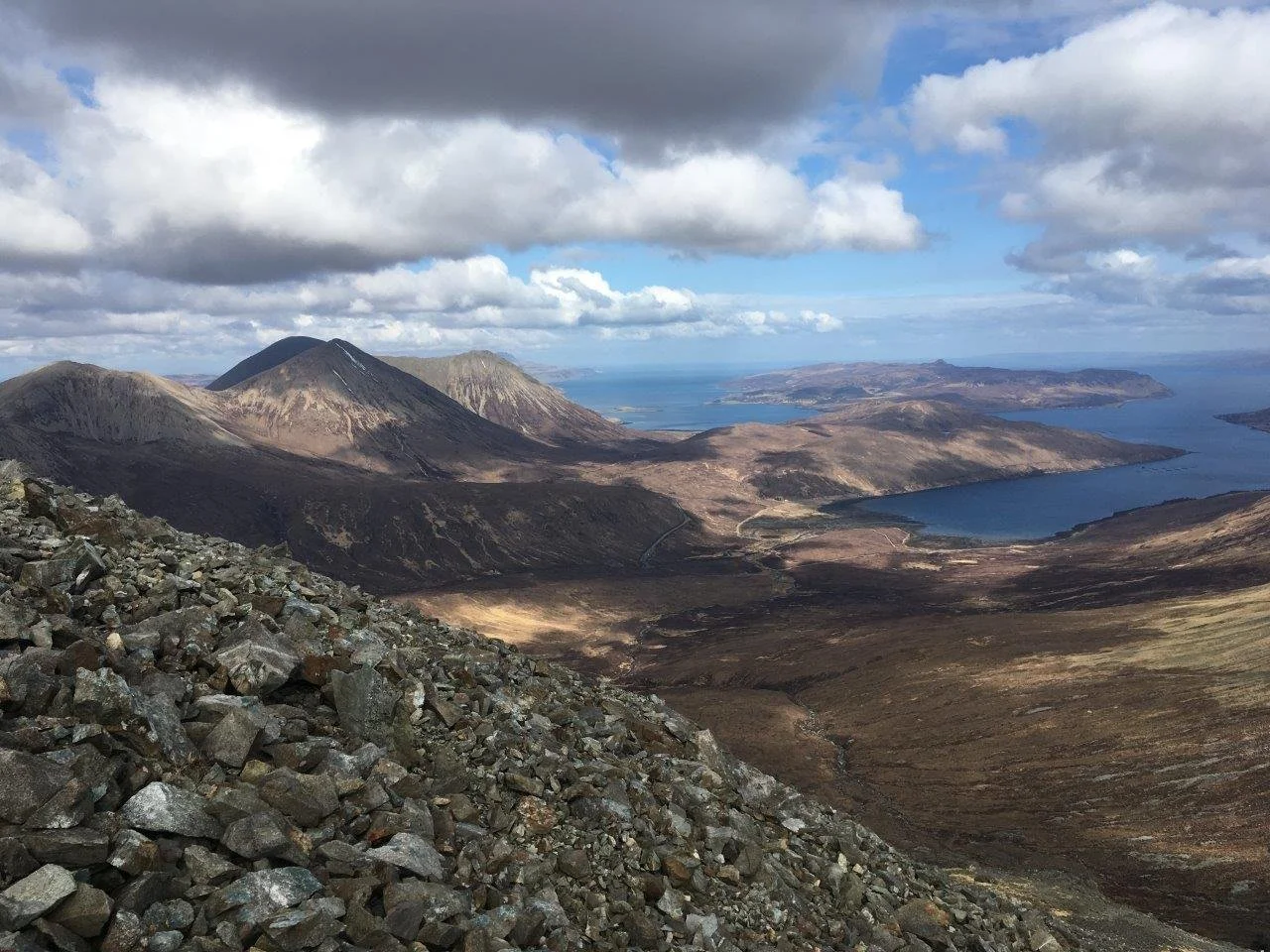 A mountainous landscape with rocky foreground, volcanic peaks, lakes, and cloudy sky.