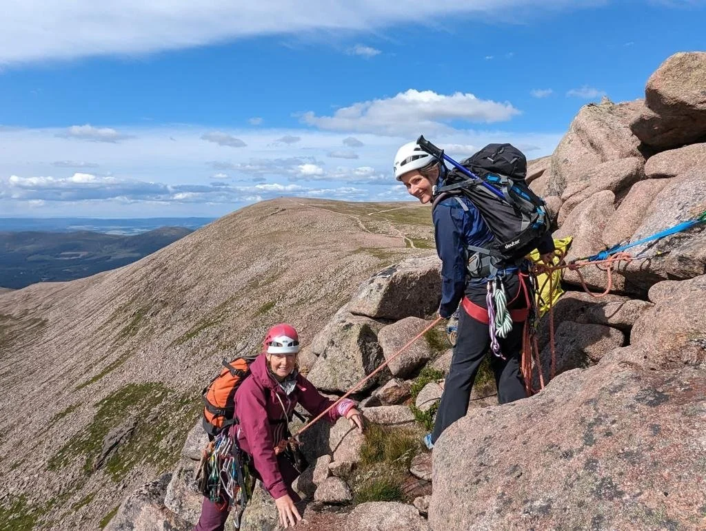 Two women in climbing gear ascending a rocky mountain trail on a clear day.