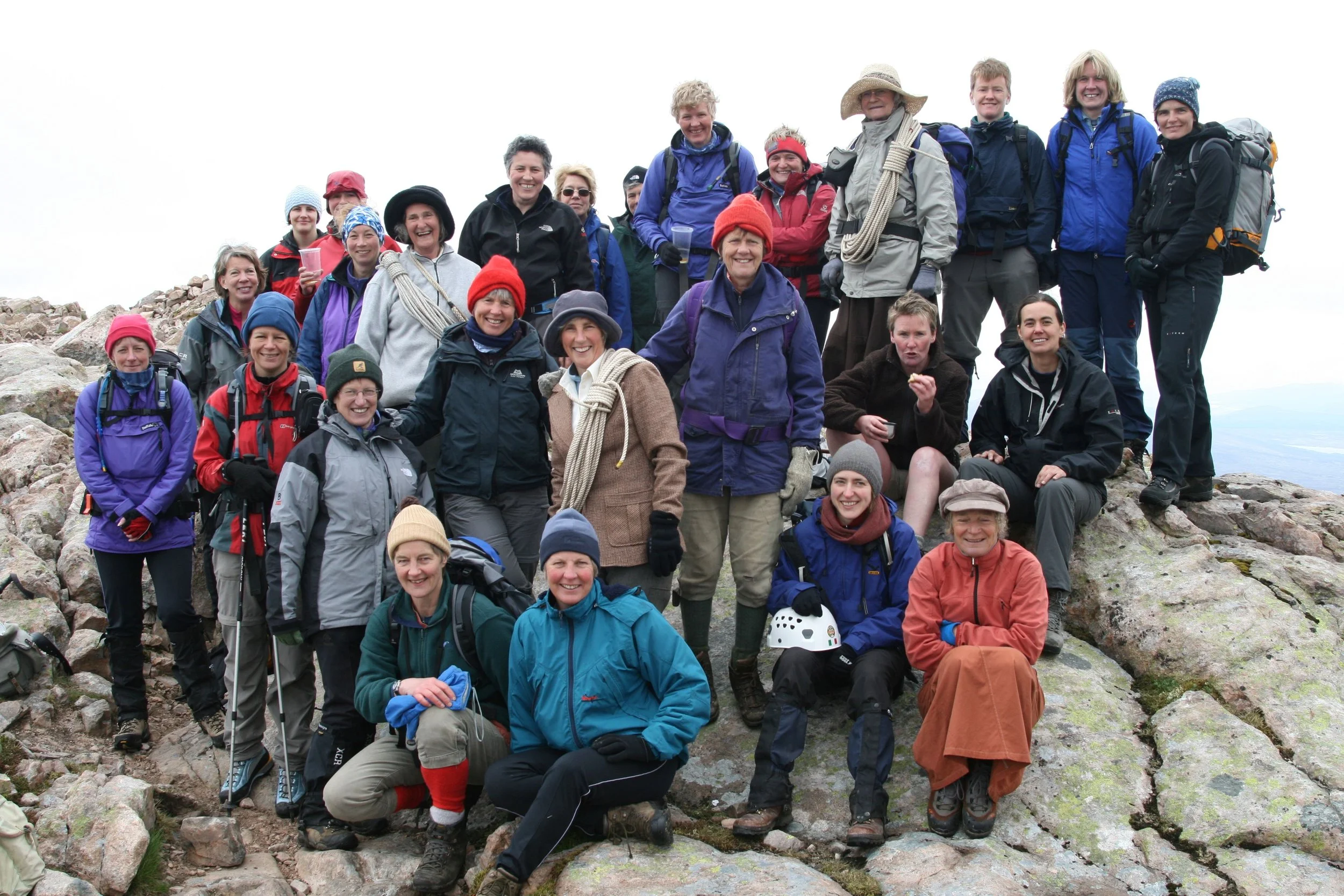 A group of hikers on a rocky mountain summit, smiling and dressed in outdoor gear. They have backpacks, hats, and weather-appropriate clothing, with some holding trekking poles and wearing helmets.