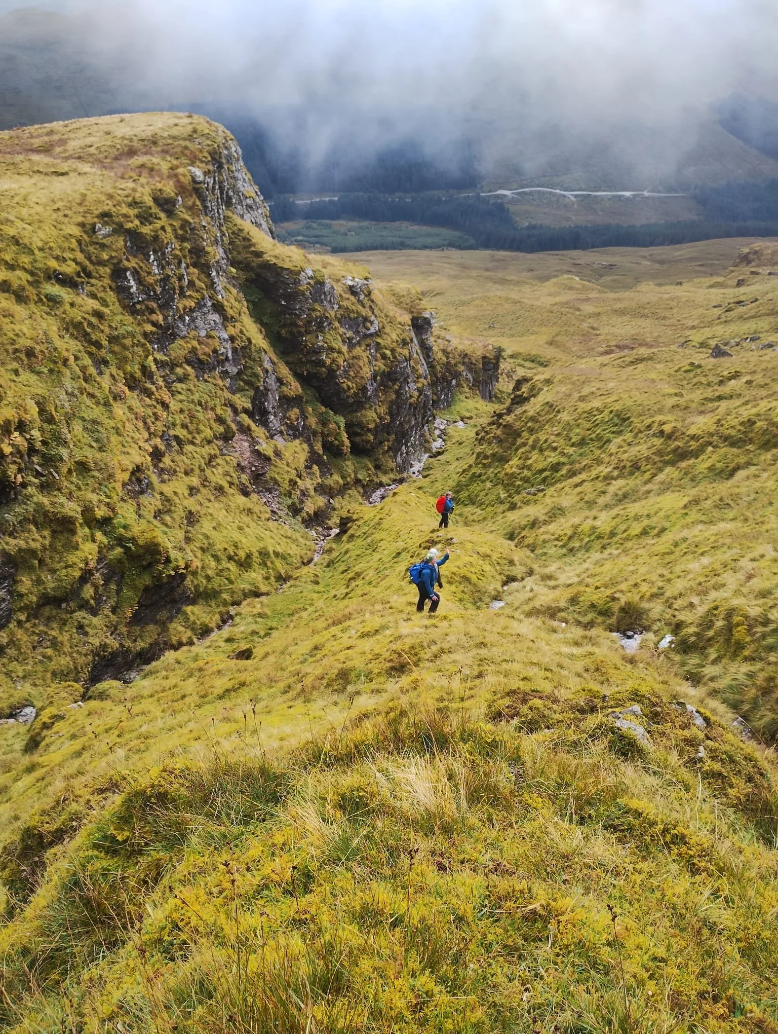 Two hikers with backpacks walking through a lush green valley surrounded by rolling hills and rugged cliffs, with a misty mountain backdrop.