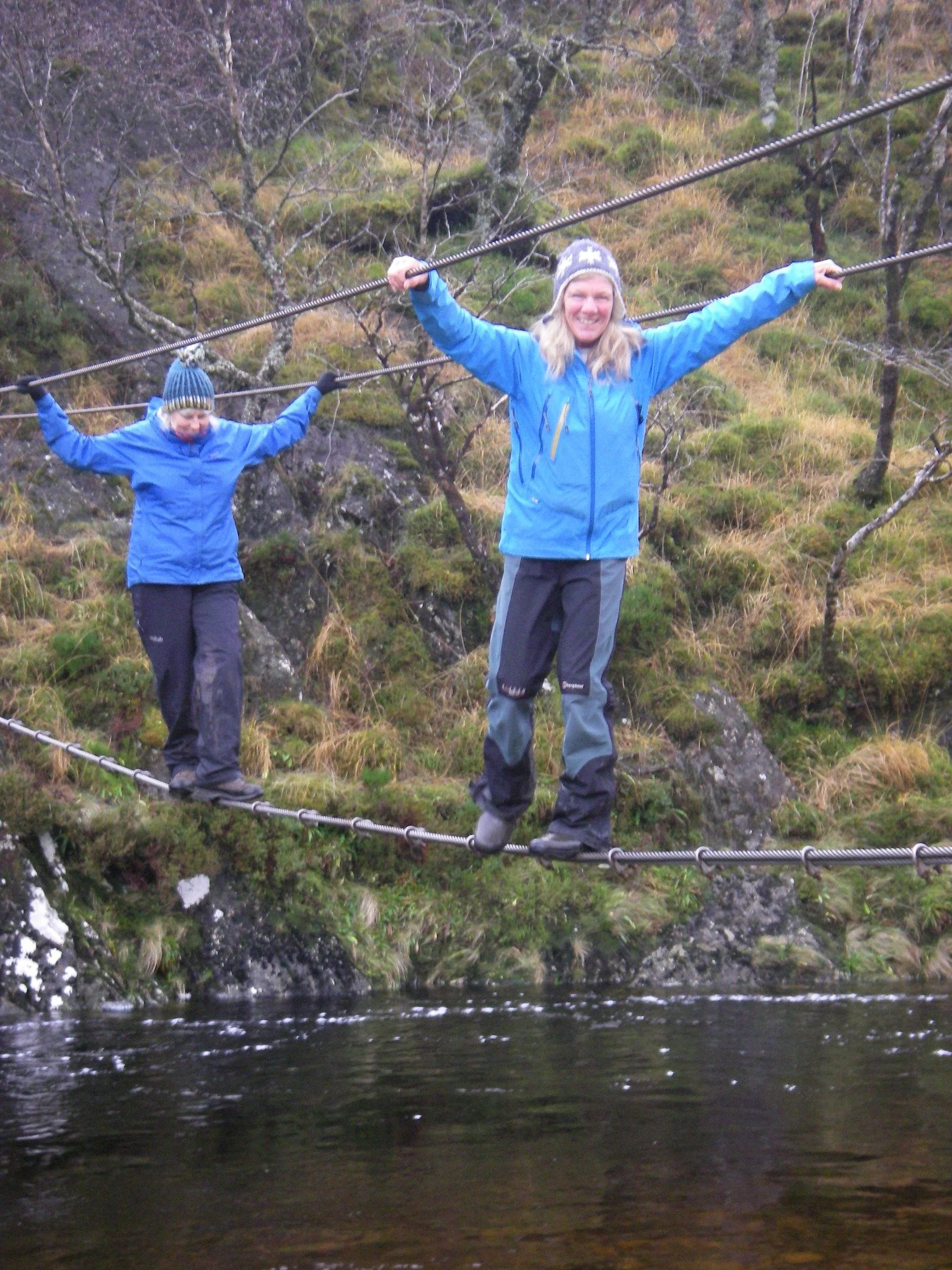 Two people crossing a narrow suspension bridge over a river, surrounded by grassy and wooded terrain, dressed in outdoor clothing and knit hats.