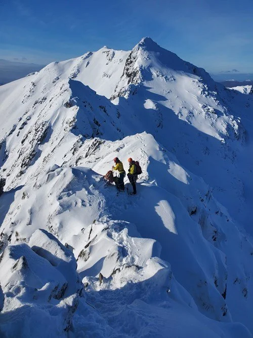 Two climbers in yellow and purple jackets on a snowy mountain ridge with a high peak in the background.
