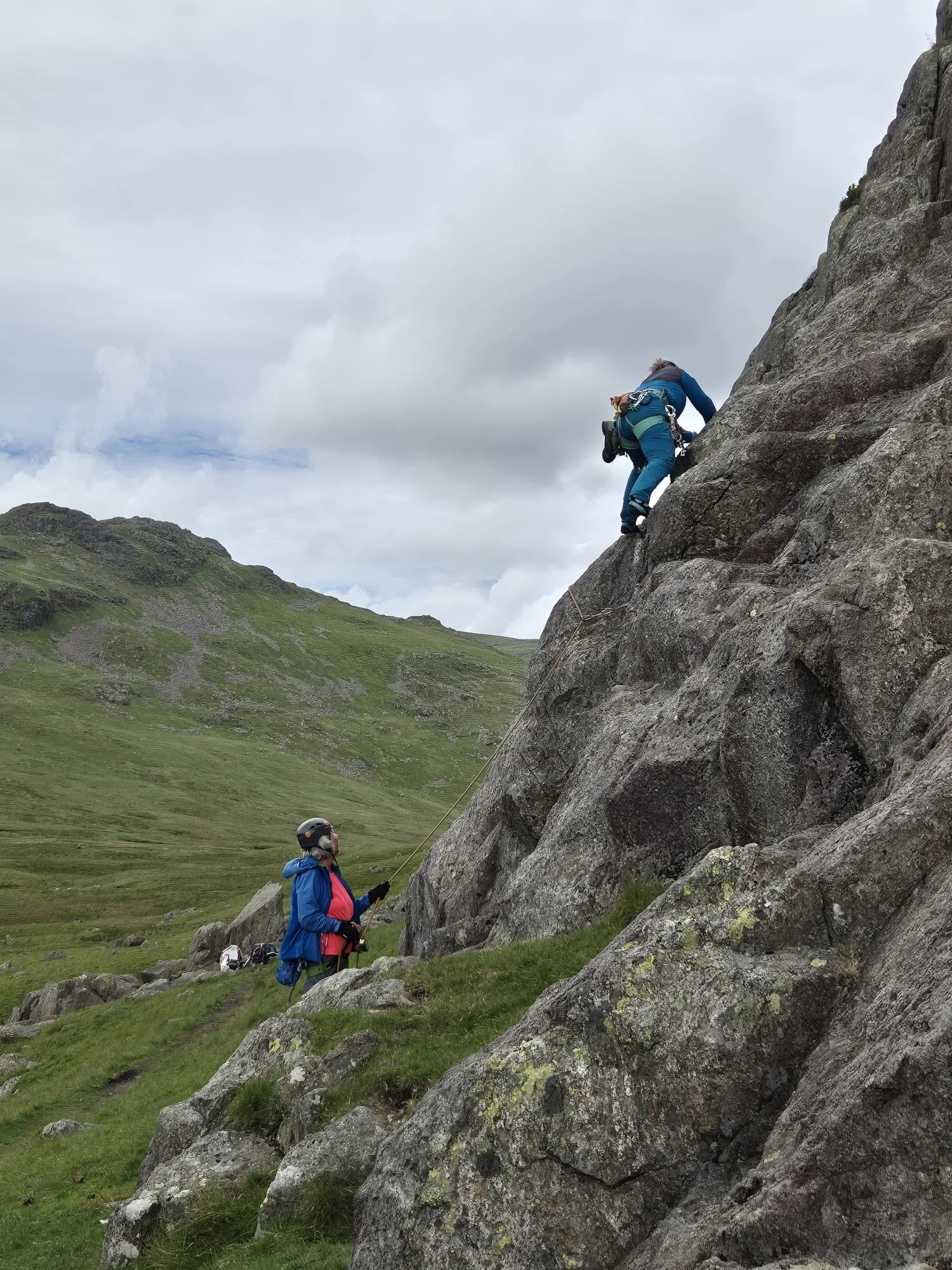 Two climbers, wearing helmets and harnesses, are ascending a large rock face in a scenic mountainous area with green grassy slopes and cloudy skies.
