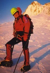 A person dressed in red mountain climbing gear, wearing a yellow helmet, standing on snow with a mountain in the background.