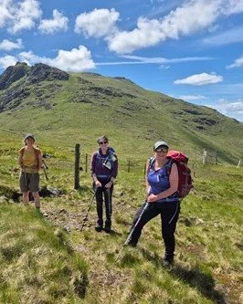 Three hikers standing on a grassy trail with a mountain in the background, blue sky, and clouds overhead.