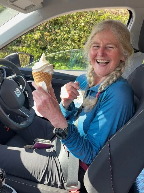 An elderly woman with long gray hair in braids, smiling and sitting in the driver's seat of a car, holding an ice cream cone. She is wearing a blue jacket, with trees and greenery visible outside the car window.