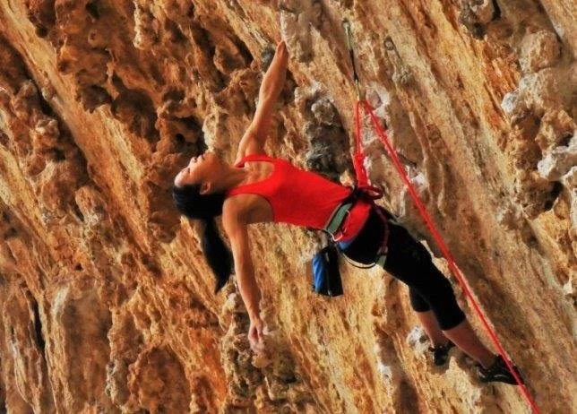 A woman rock climbing on an indoor or outdoor rock face, wearing a red tank top, black pants, and climbing gear, including a harness and safety rope.