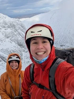 Two people in outdoor winter gear smiling on a snowy mountain with glacier ice in the background.