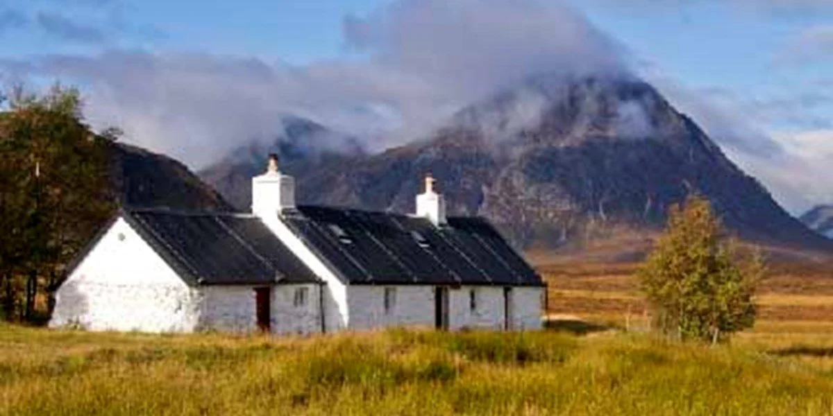 White houses with black roofs in a grassy field, mountain range in the background with clouds above.
