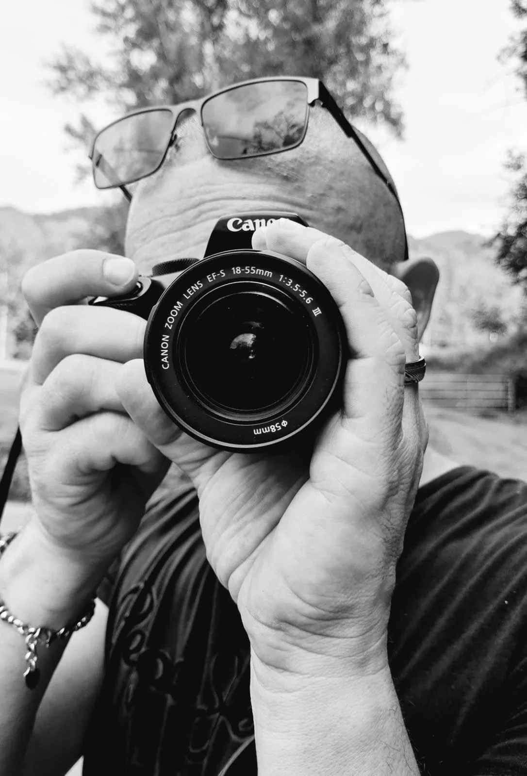 Un homme porte des lunettes et tient un appareil photo Canon noir devant son visage, dans un environnement extérieur. Il prend une photo en regardant à travers l'objectif.