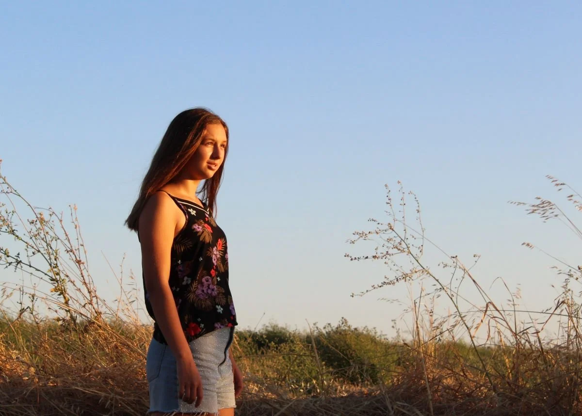 Jeune femme debout dans une prairie, vêtue d'un haut noir à motifs floraux et d'un short en jean, regardant à l'horizon sous un ciel clair.