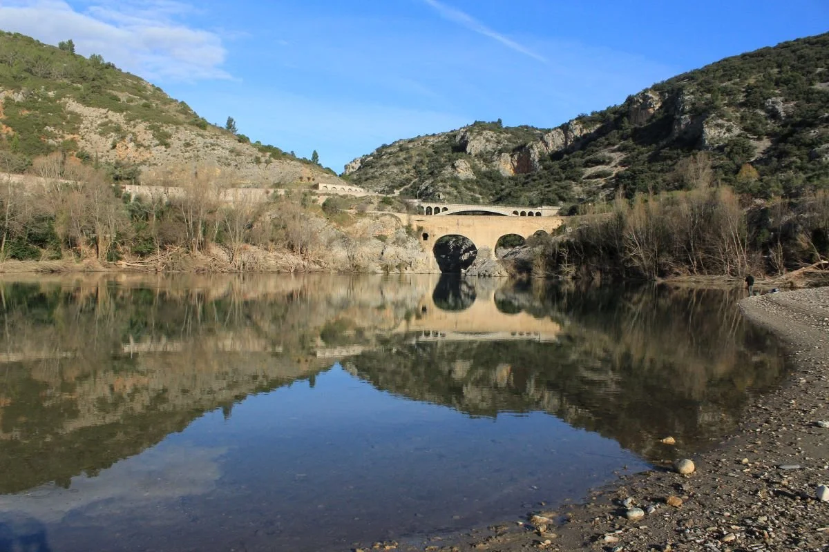 Paysage avec une rivière reflétant un pont en pierre dans une vallée entourée de montagnes et d'arbres.