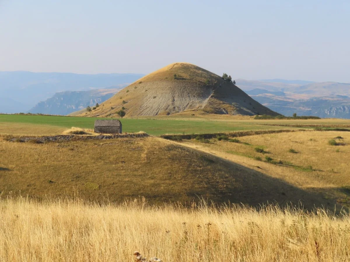 Un paysage avec une grande colline ou montagne ronde, des champs verts et dorés, un petit bâtiment en pierre, et un ciel clair.
