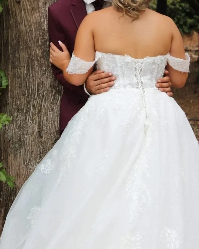 Deux personnes habillées en vêtements de mariage, l'une en robe blanche de mariée et l'autre en costume marron, debout près d'un arbre.