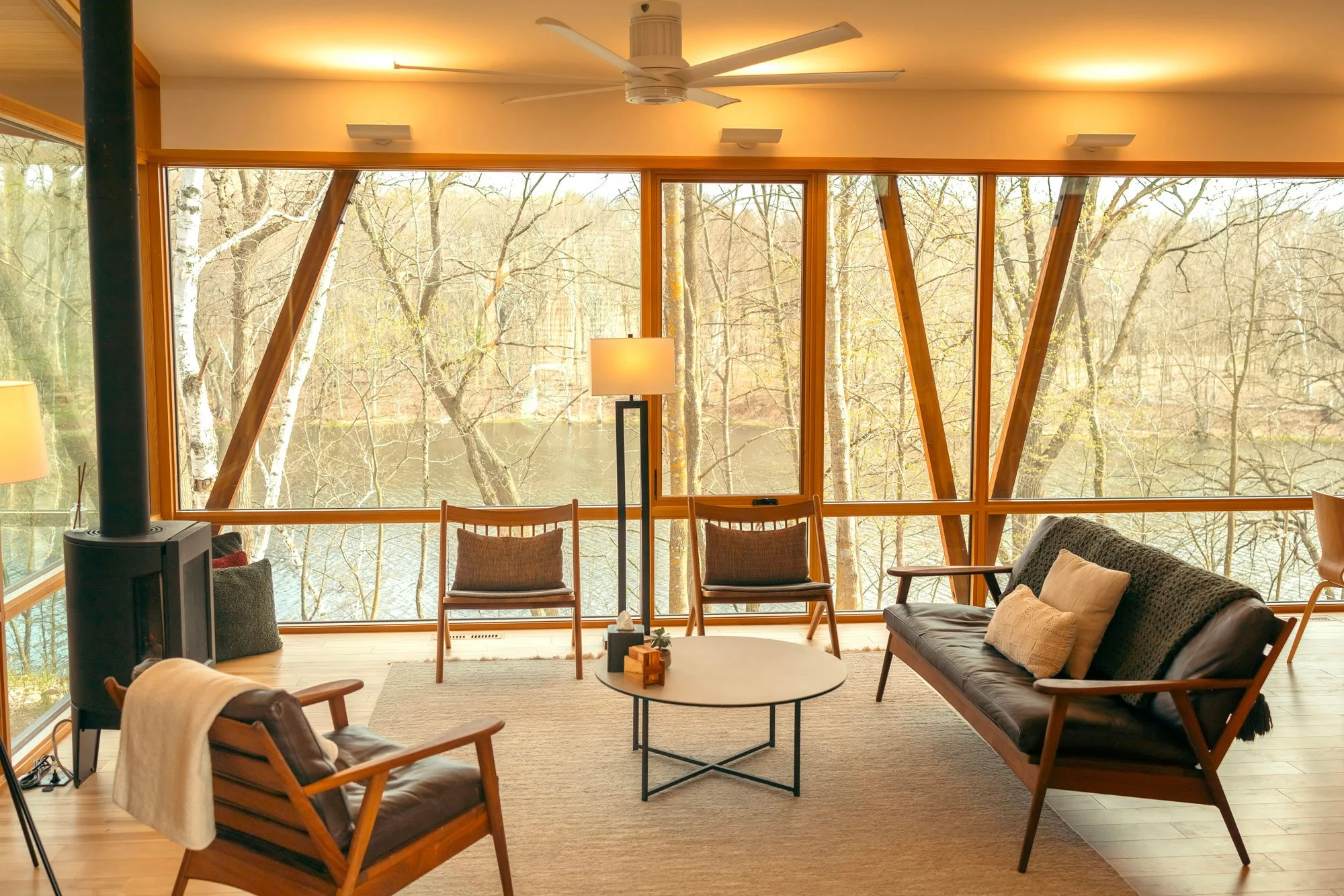 Salon avec grandes fenêtres donnant sur une forêt, mobilier en bois et fauteuils en cuir, lampes et ventilateur au plafond.