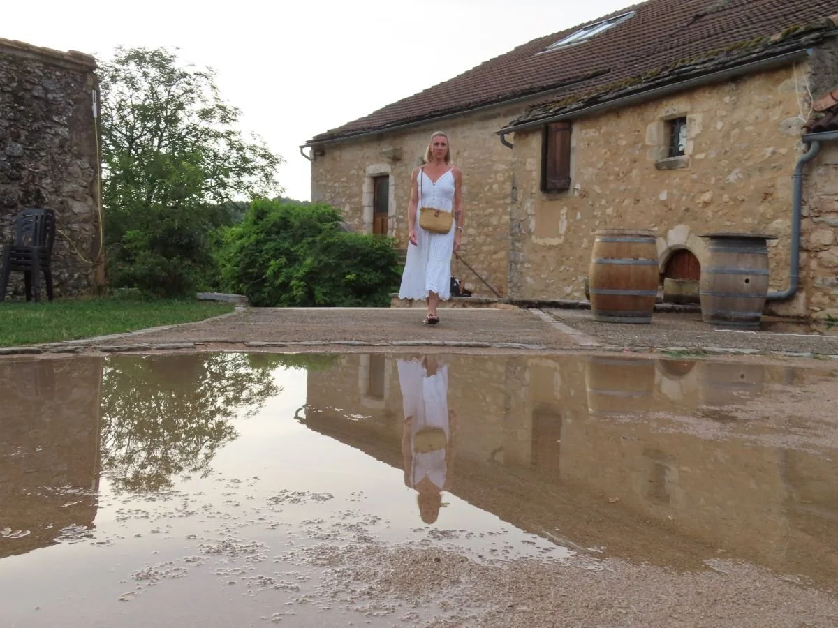 Une femme en robe blanche marche sur une terrasse en pierre, dont le reflet est visible dans une flaque d'eau au premier plan, devant une vieille maison en pierre avec des barils en bois à côté.