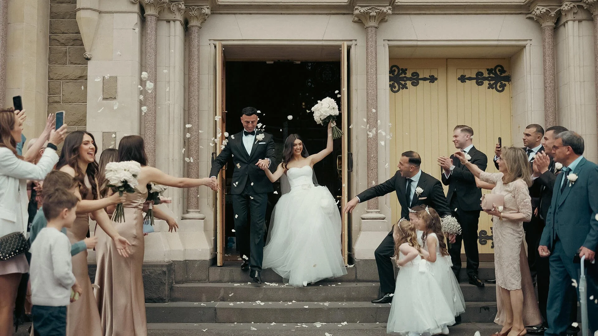 A newly married couple leaves a church, holding hands, with friends and family celebrating outside. The bride in a white gown holds a bouquet and smiles, while the groom in a tuxedo also smiles. Guests are throwing flower petals and taking photos.