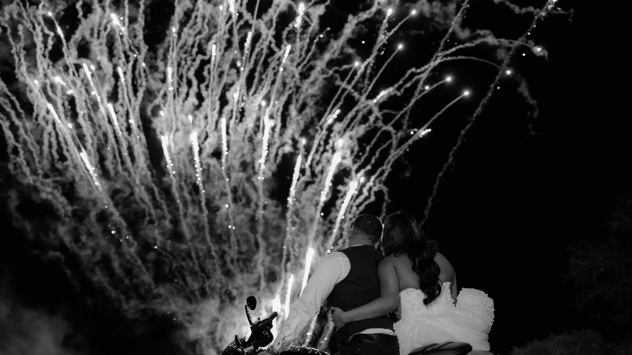 Couple watching fireworks at night, with the fireworks illuminating the sky in the background.