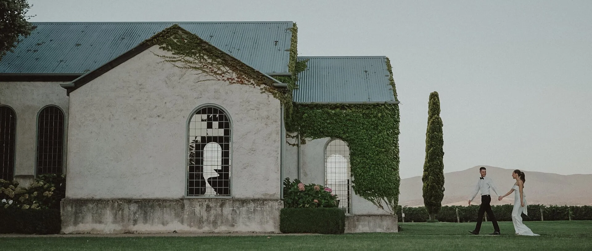A bride and groom holding hands and walking on a lawn next to a white church with ivy-covered walls and stained glass windows, against a backdrop of mountains and a pale sky.