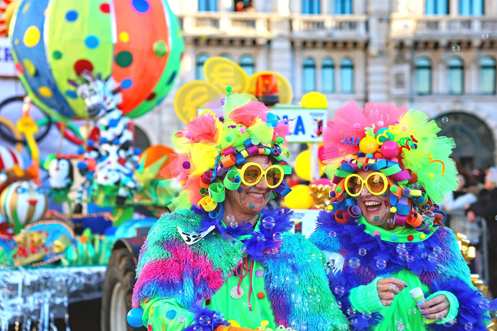 Persone vestite con costumi colorati e decorati con nastri, tulle e occhiali con forma di cuore e tondi, partecipano a una festa o parata colorata in una piazza.