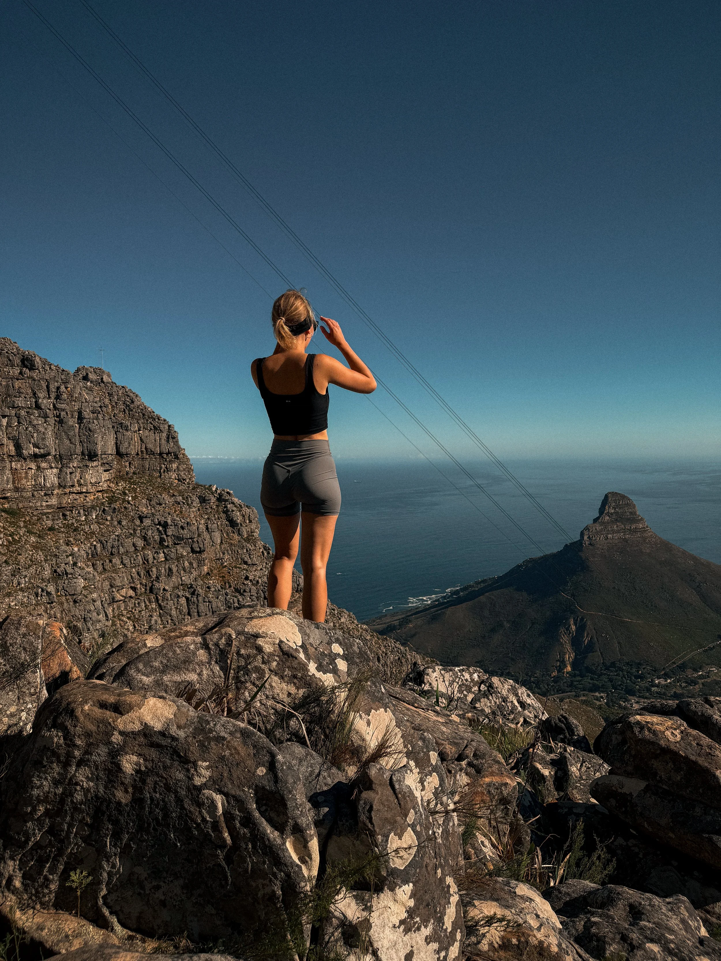 Junge Frau in Sportkleidung auf felsigem Berggipfel mit Blick auf das Meer und eine markante Felsformation im Hintergrund, bei Sonnenuntergang.